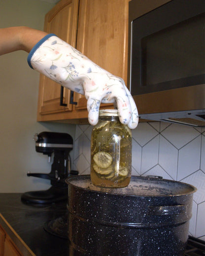 A gloved hand holding a preserving jar above a canning pot on the stove